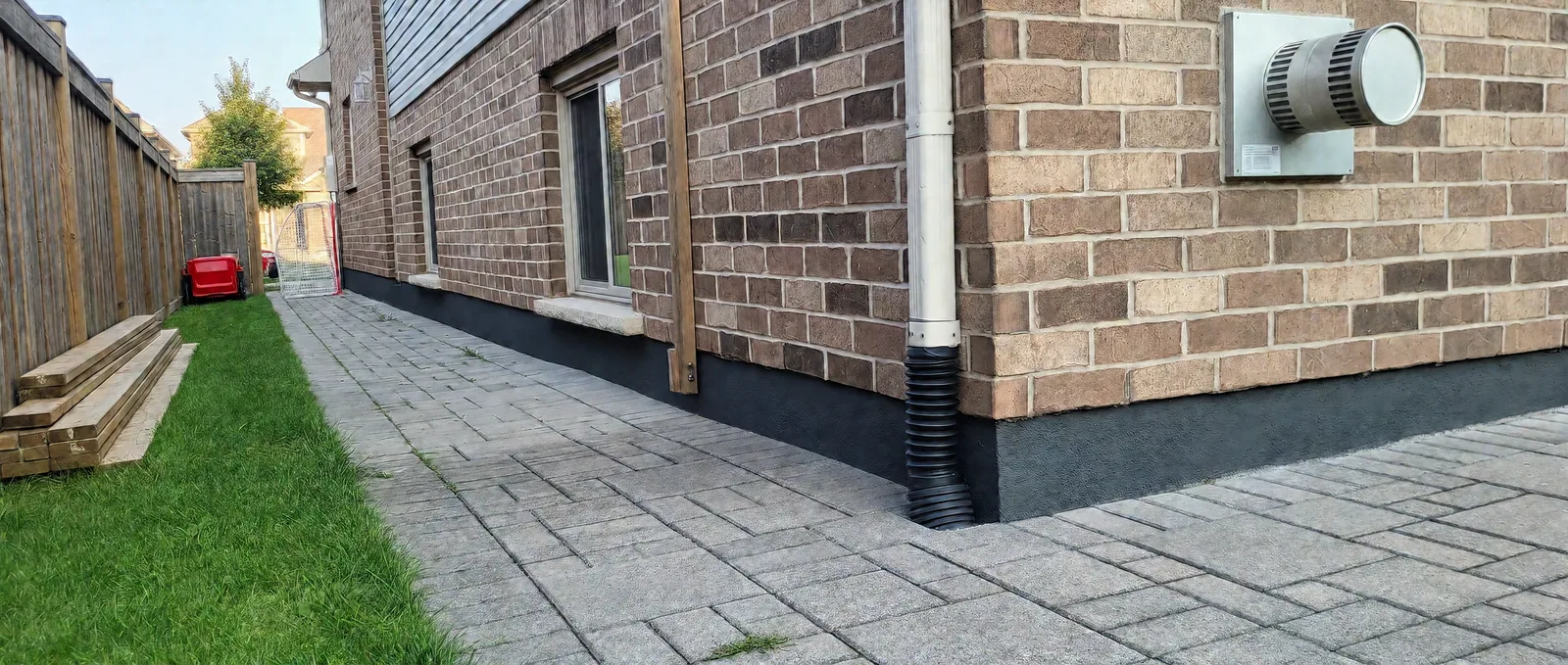 Continuous charcoal parge finish along the side of a brick home in Niagara Falls, beneath basement windows and around the corner beside a paver walkway